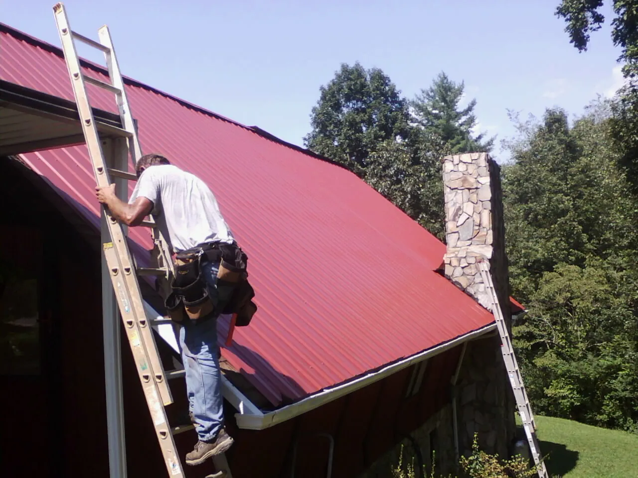 Roofer on a ladder beside a red metal roof and stone chimney — Soffit & Fascia Repair by Roofing Brothers in Dandridge